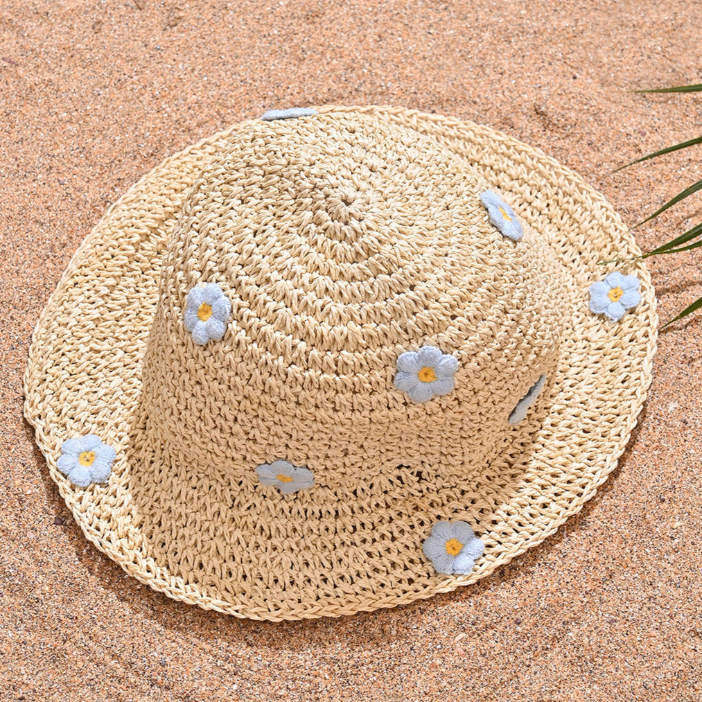 A beige woven straw summer beach hat with blue floral accents, displayed on a sandy surface with a blue water background.