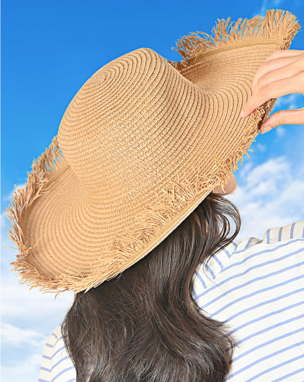 A woman wearing a tan foldable visor hat with a wide brim, held up to her head against a blue sky background, suggesting a summer beach setting.