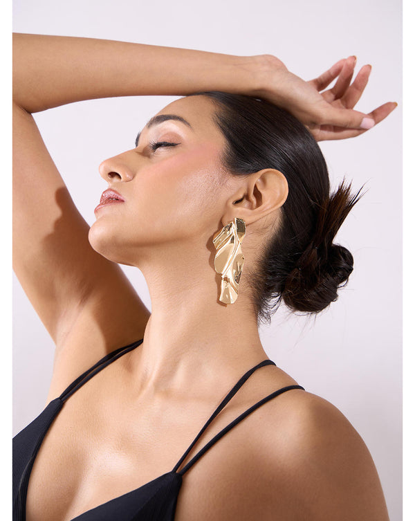 A woman wearing gold molten leaf drop and dangler earrings with a black top, against a white background.