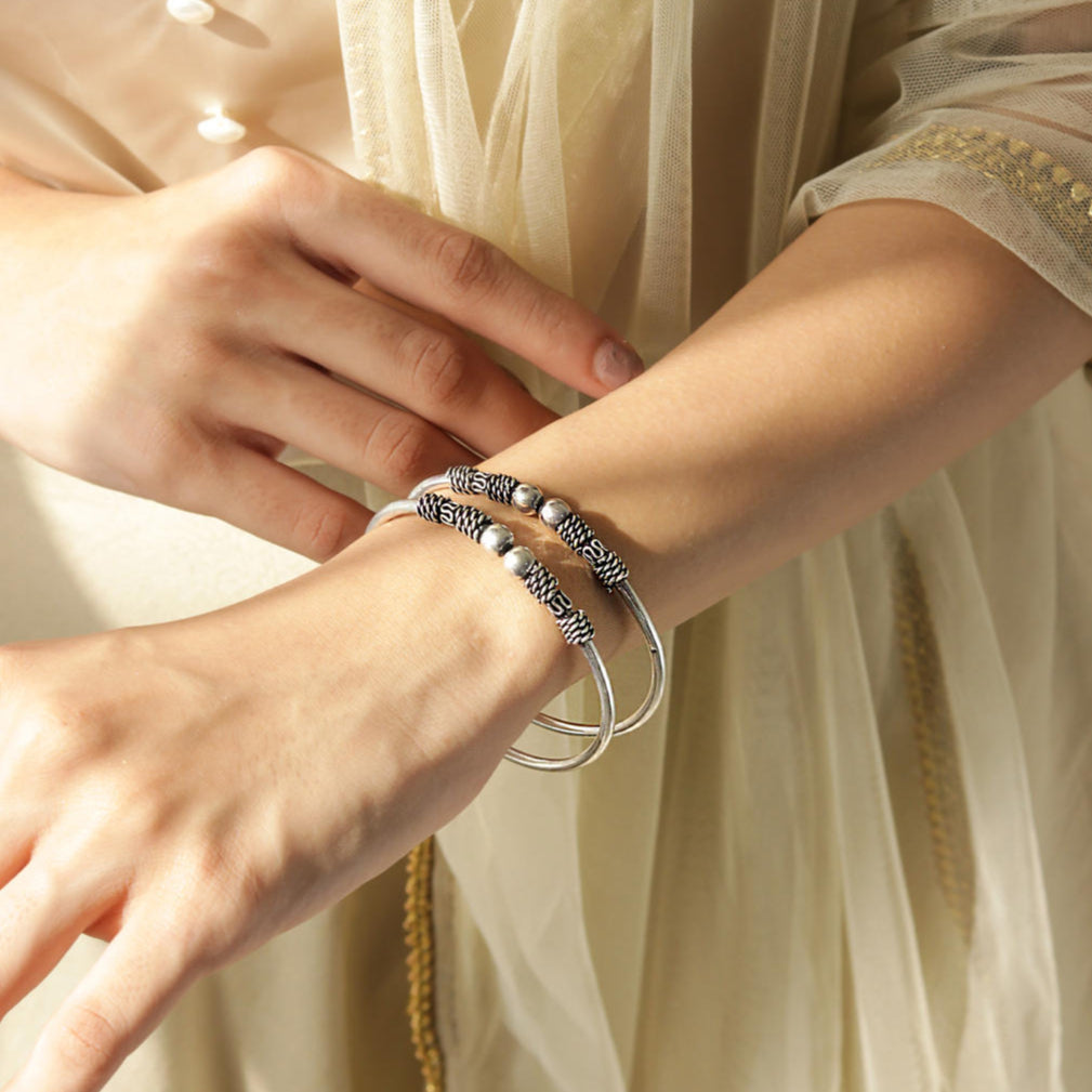 A silver-plated oxidised bangle set worn on a person's wrist, featuring intricate detailing and a textured look.
