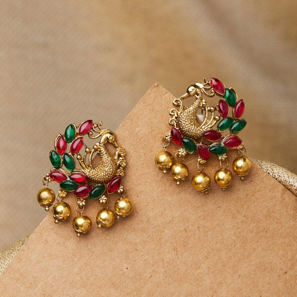 A pair of gold-plated peacock stud earrings with red and green stone embellishments on a beige surface.
