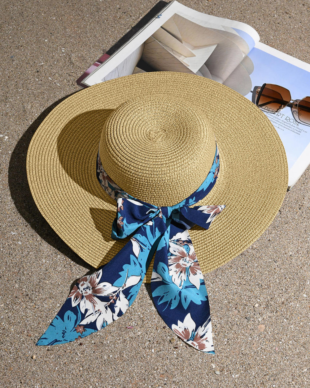 A straw beach hat with a navy printed scarf around the brim, displayed on a surface near a pair of sunglasses and a magazine.