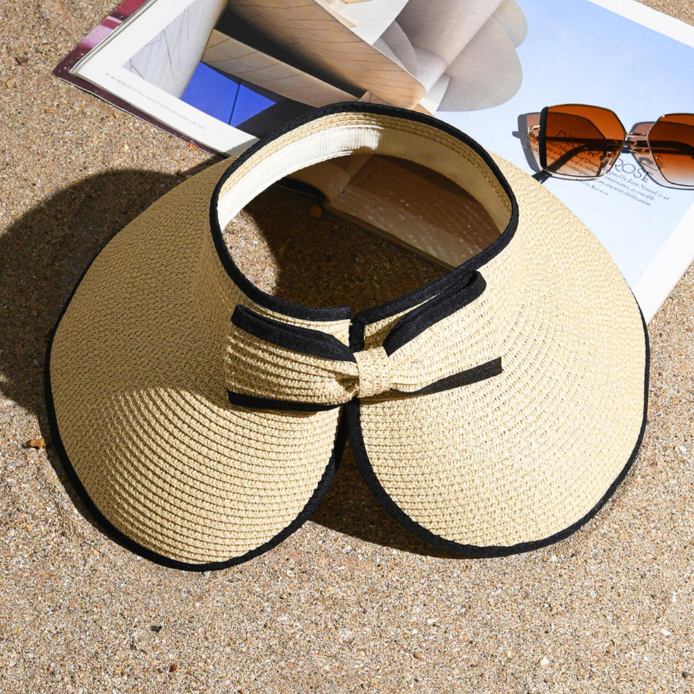 A beige sun visor hat with black edging and a bow at the back, displayed on a sandy surface with a pair of sunglasses and a book in the background.
