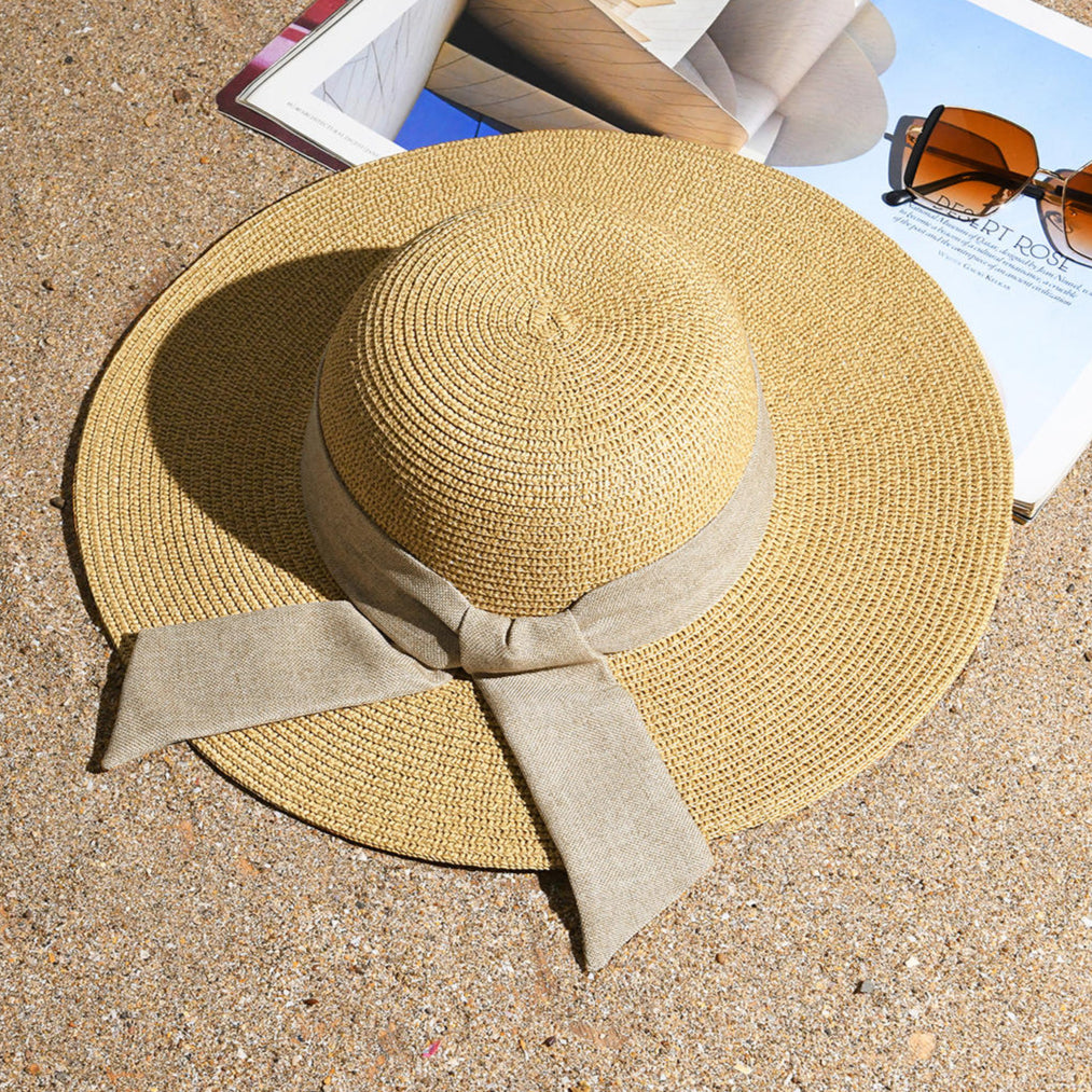 A beige beach hat with a khaki scarf tied around the base, placed on a sandy surface with a magazine and sunglasses in the background.