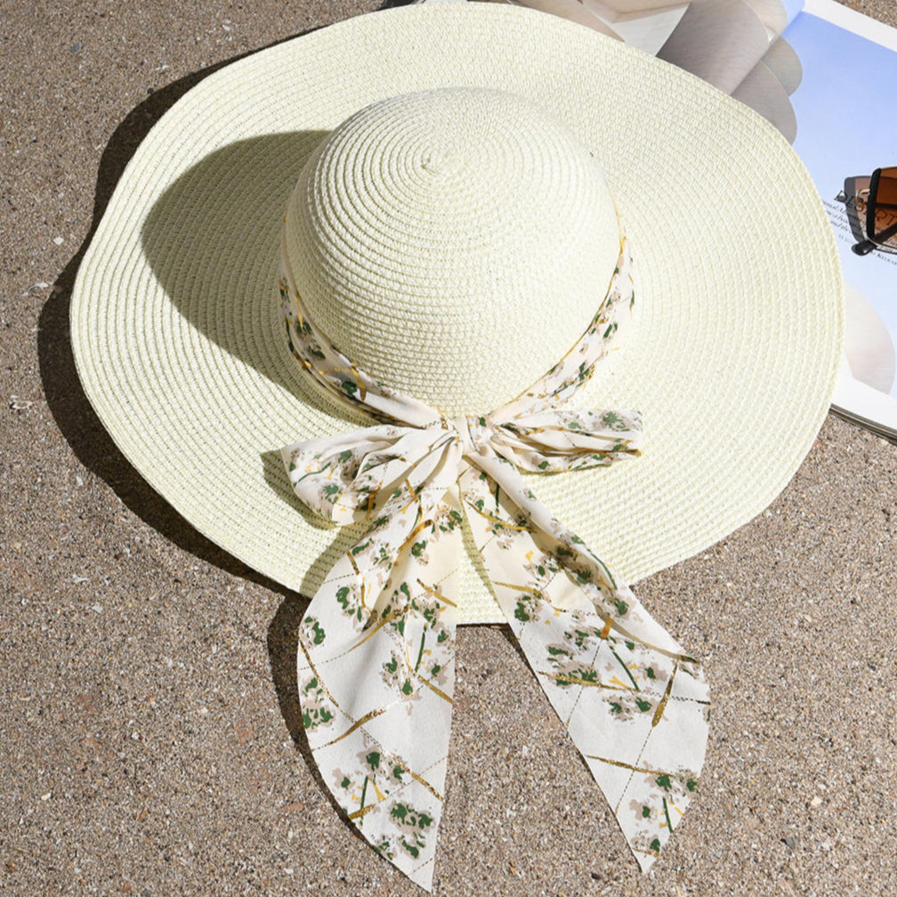 A white beach hat with an ivory floral scarf tied around the brim, placed on a sandy surface with a pair of sunglasses and a book in the background.