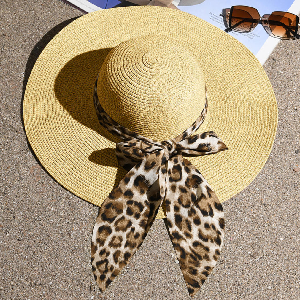 A beige beach hat with a leopard print scarf tied around the base, placed on a surface with a magazine and sunglasses in the background.