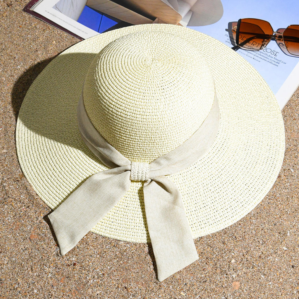 A white beach hat with a tan scarf tied around the base, placed on a sandy surface with a magazine and sunglasses in the background.