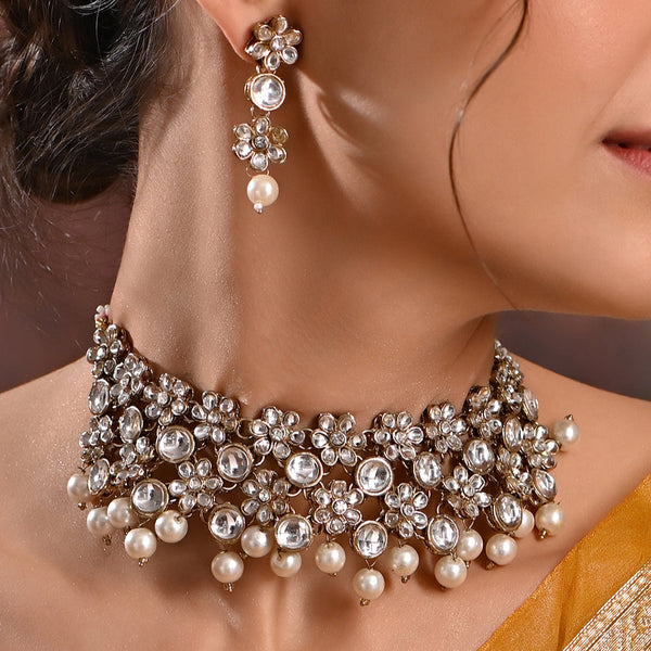A close-up image of a gold-plated floral jewellery set with kundan and pearl accents, consisting of a choker necklace and a pair of jhumki earrings.