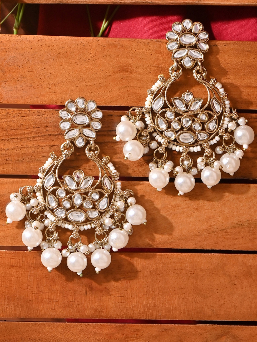 A pair of gold-toned chandbali earrings with white kundan stones and ivory pearl drops, displayed against a wooden background. 