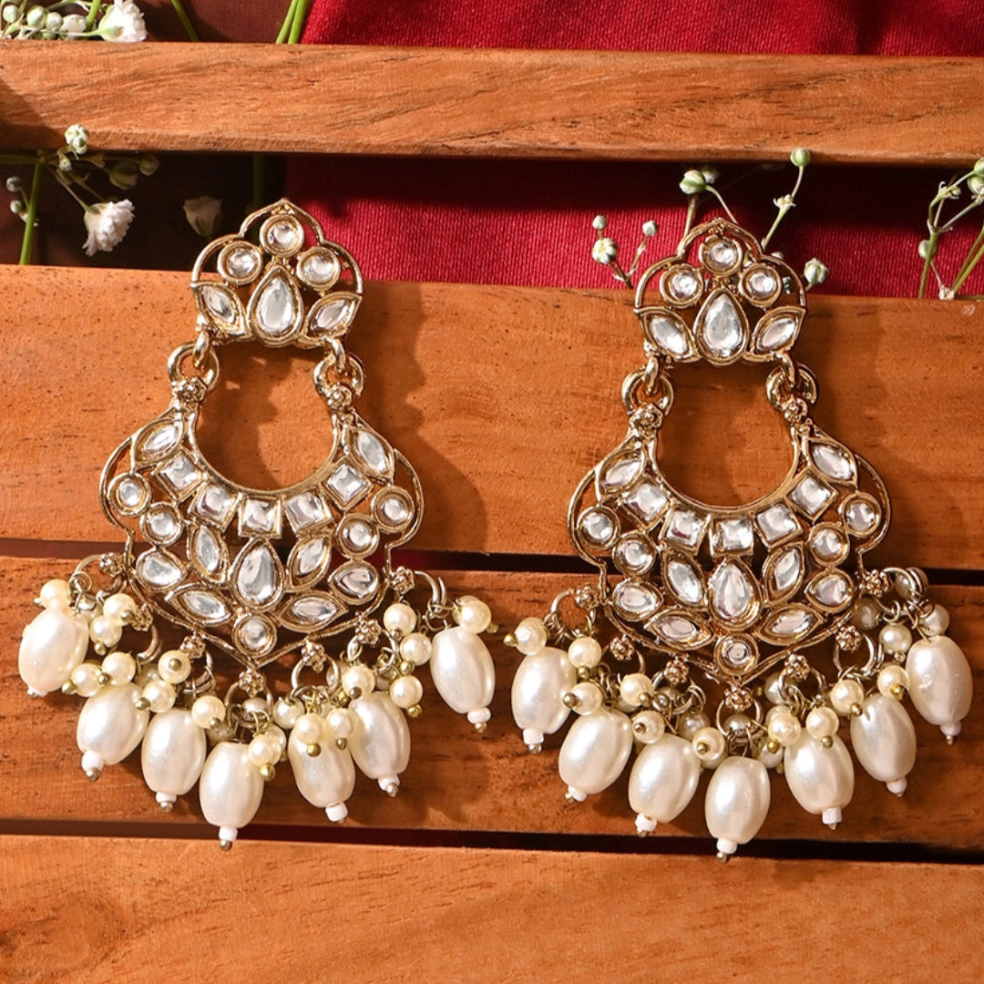 A pair of gold-plated chandbali earrings adorned with white kundan stones and pearl drops, displayed against a wooden background with floral embellishments.