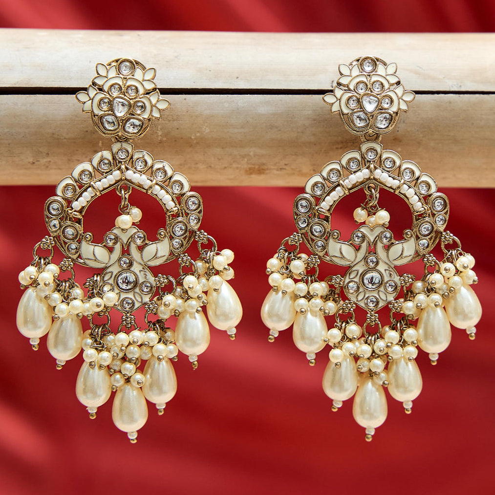 A pair of gold-toned Chandbali earrings with white Meenakari detailing and pearl drops, displayed against a red background.