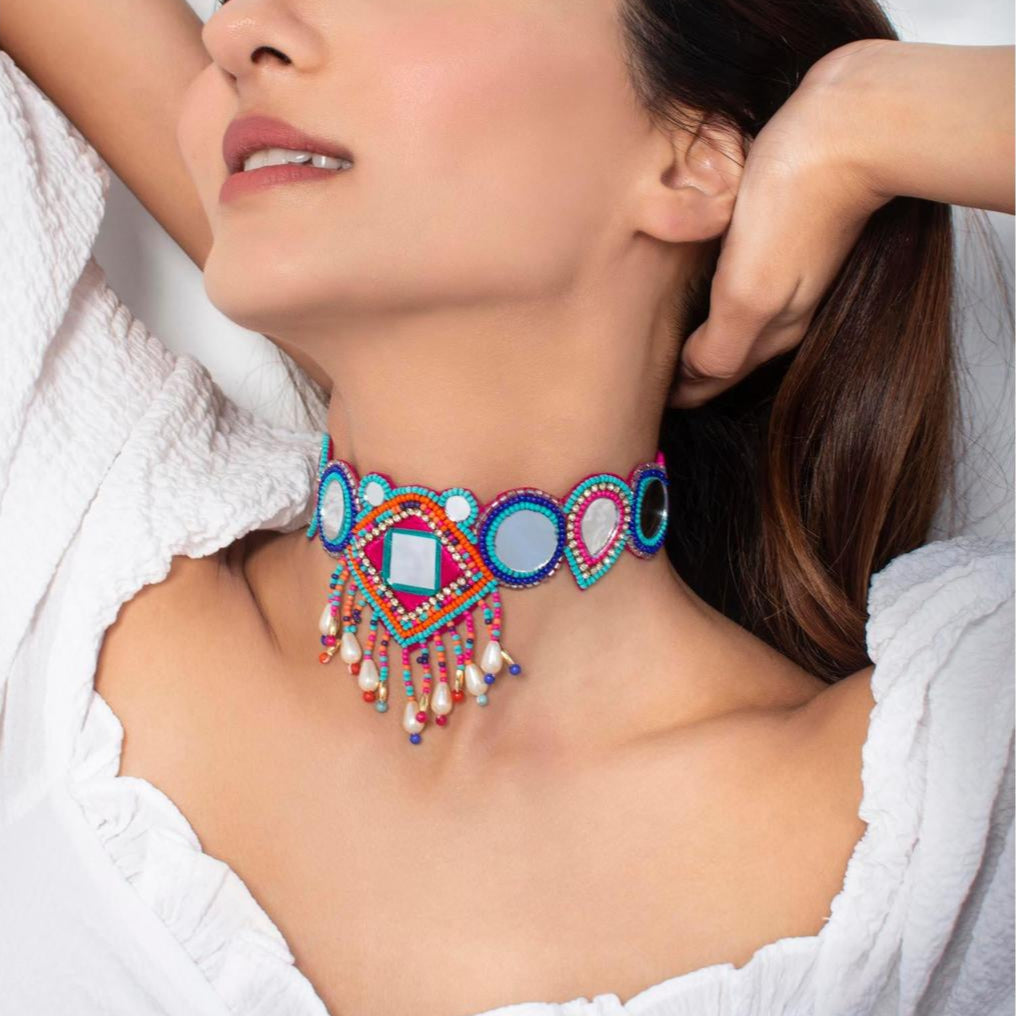 A person wearing a white outfit showcasing a multicolor beaded choker necklace with geometric patterns and mirror accents.
