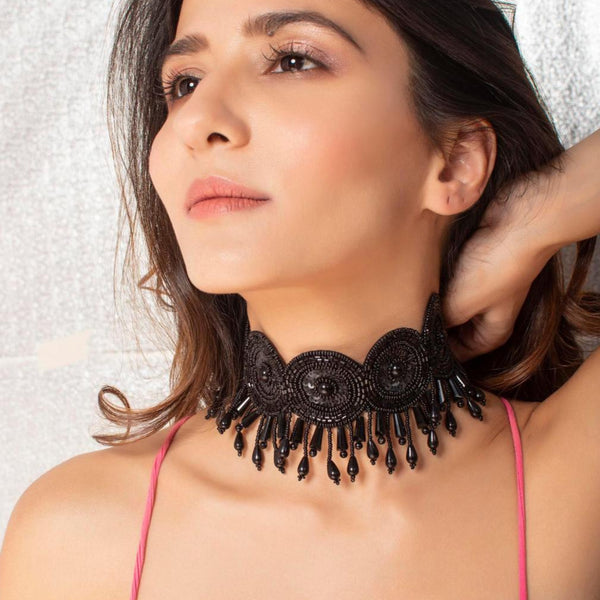A woman wearing a pink top and showcasing a black seed bead choker necklace with floral motifs and pearl accents.