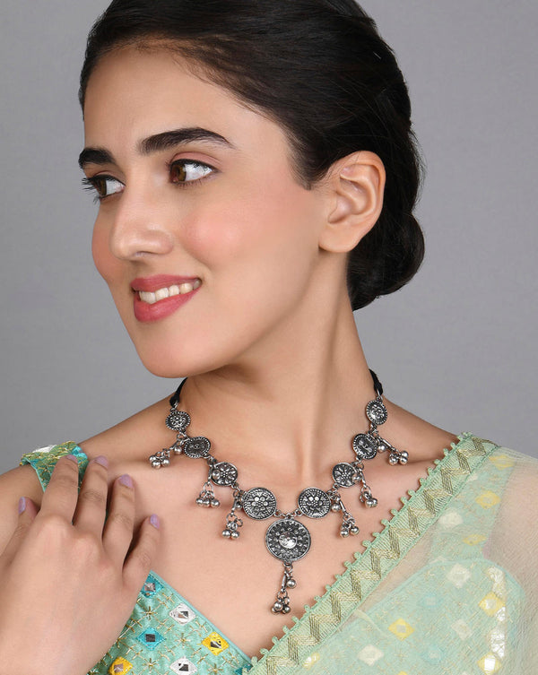 A woman wearing a Patina Coin Choker Necklace with an oxidized silver finish and coin motifs, paired with a light green saree with golden floral patterns.