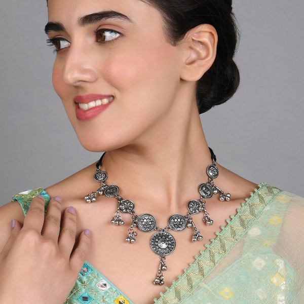 A woman wearing a Patina Coin Choker Necklace with an oxidized silver finish and coin motifs, paired with a light green saree with golden floral patterns.