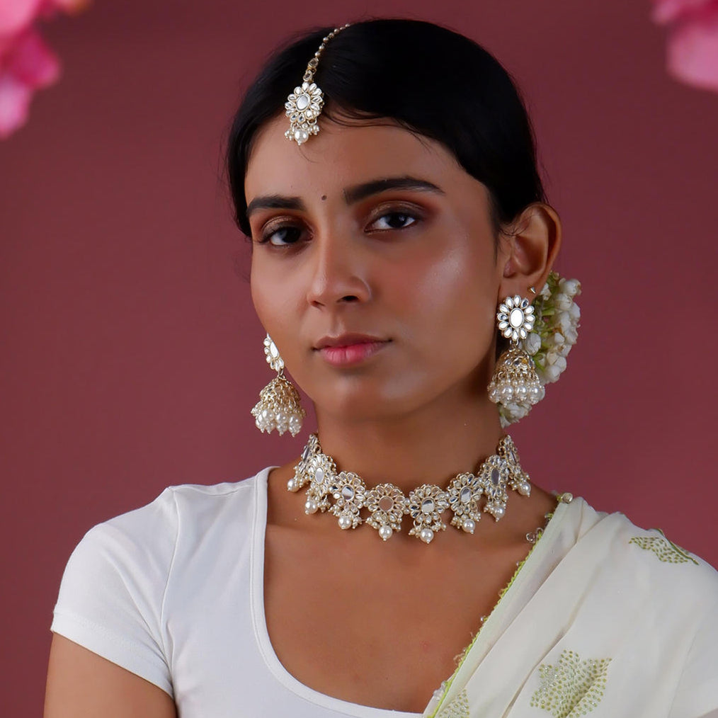 A woman wearing a traditional pearl and mirror kundan jewellery set which includes a choker necklace, earrings, and a maang tikka. The set is gold-plated with white artificial stones.