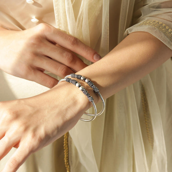 A silver-plated oxidised bangle set worn on a person's wrist, featuring intricate detailing and a textured look.