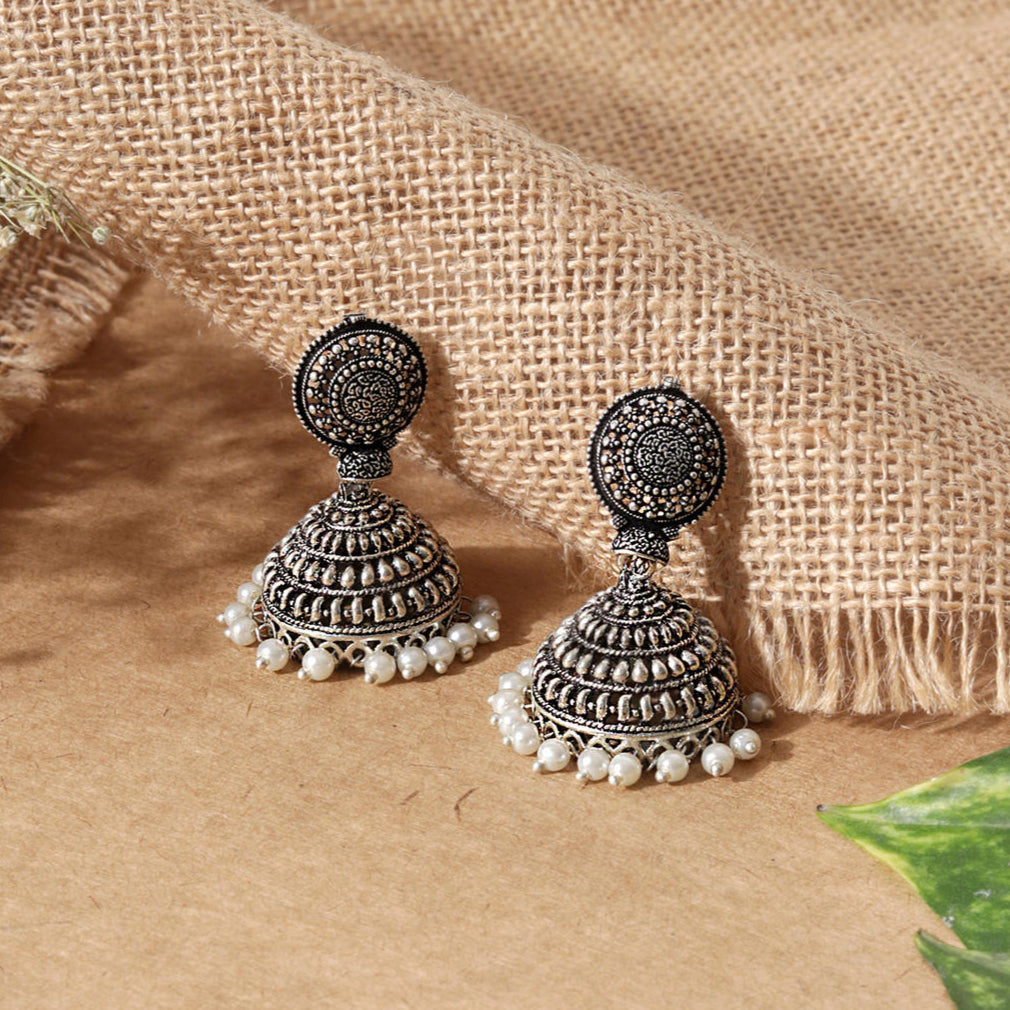 Two silver-plated oxidised jhumka earrings with intricate designs and small pearl embellishments, displayed against a burlap fabric background.