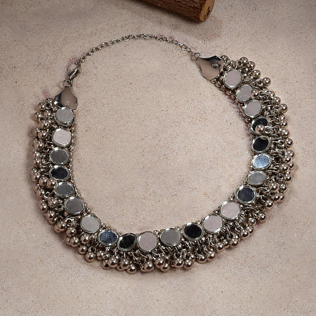 A silver-plated oxidised choker necklace with mirror work and tiny ghungroos, displayed against a beige background.