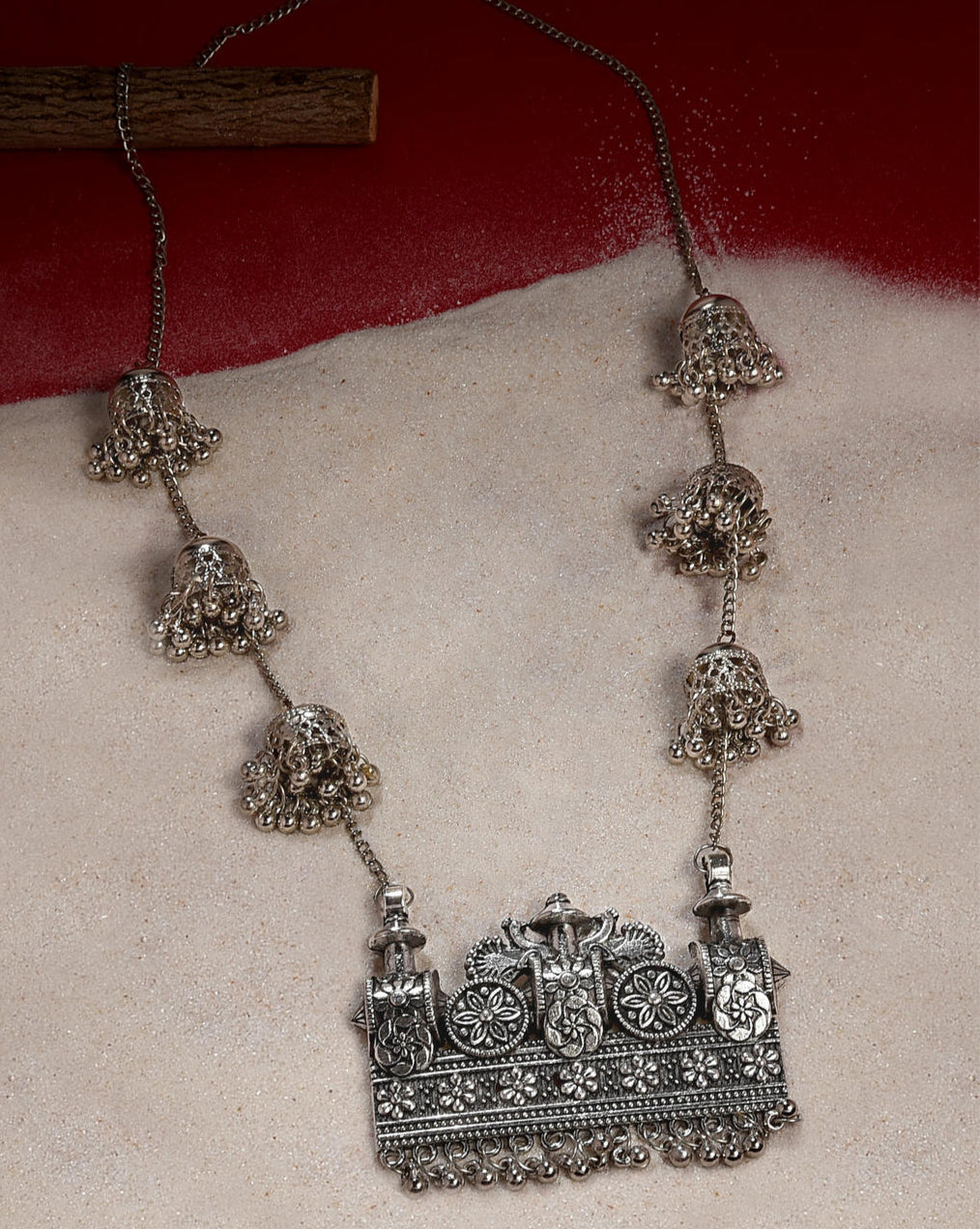 An oxidised silver long necklace with intricate chariot-inspired patterns and delicate jingles, displayed against a red and white background.