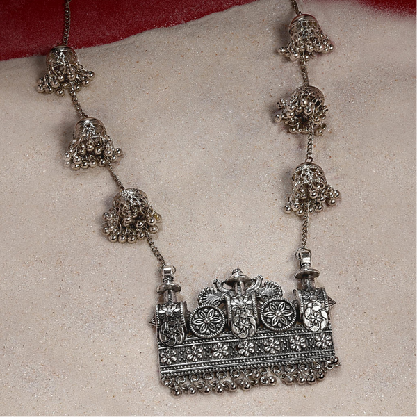 An oxidised silver long necklace with intricate chariot-inspired patterns and delicate jingles, displayed against a red and white background.