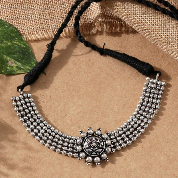 A silver oxidised choker necklace with a circular mandala-inspired pendant at the center, suspended from a black thread chain on a beige background with a green leaf to the side.
