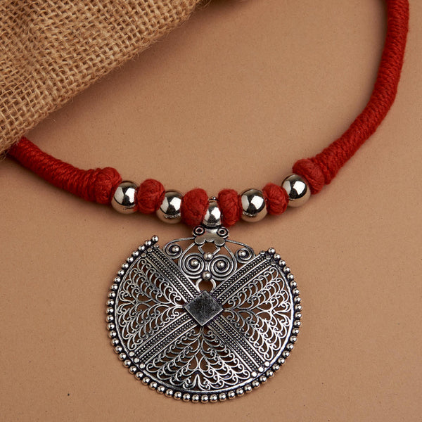 An oxidized mandala pendant necklace with intricate detailing, attached to a red thread with silver beads, presented on a beige background with a burlap fabric texture.