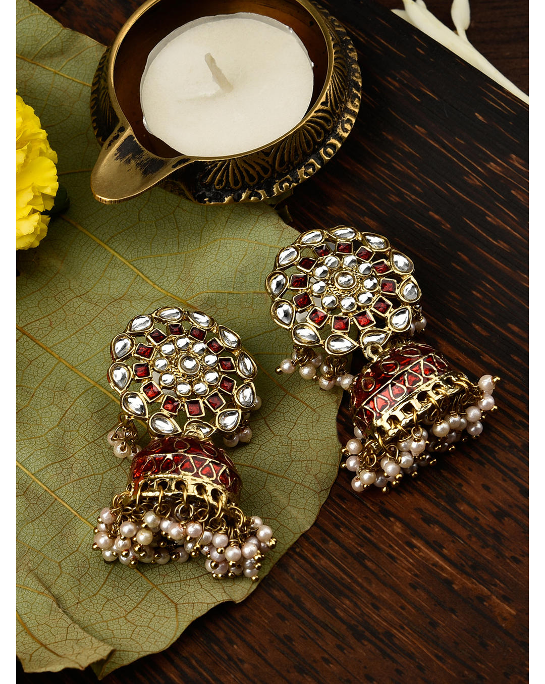 Gold-plated maroon enamelled kundan bead jhumka earrings with a spherical design, displayed on a wooden surface with a leaf and a candle.