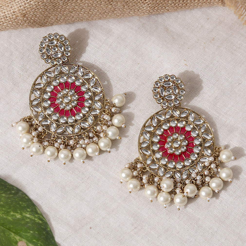 A pair of gold-plated earrings with fuchsia enamel and white pearl embellishments, displayed against a neutral background with a green leaf.
