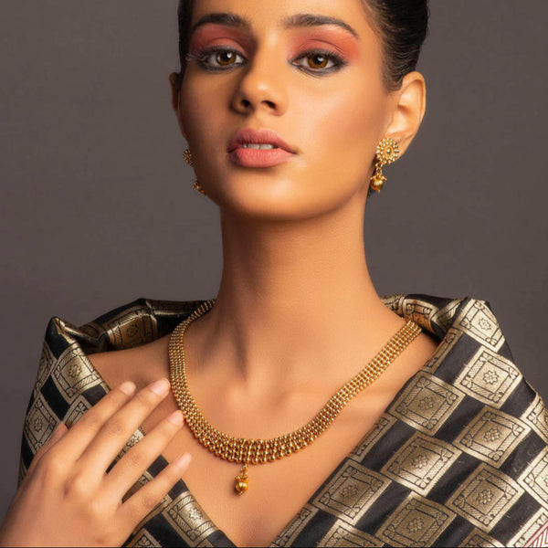 A woman wearing a gold-plated metal beaded statement jewellery set, including earrings and a necklace, with a traditional checkered garment.