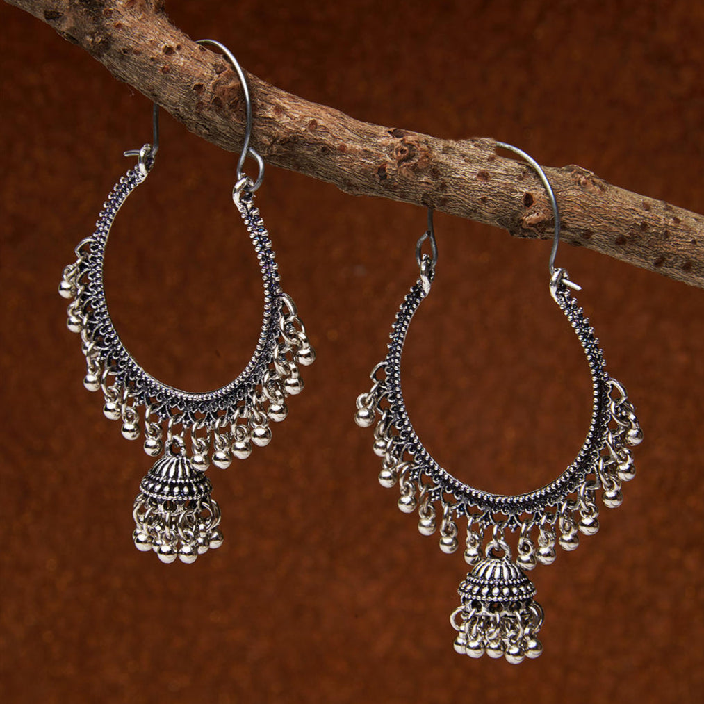 A pair of silver-plated oxidised oversized hoop jhumka earrings displayed on a wooden branch against a brown background.