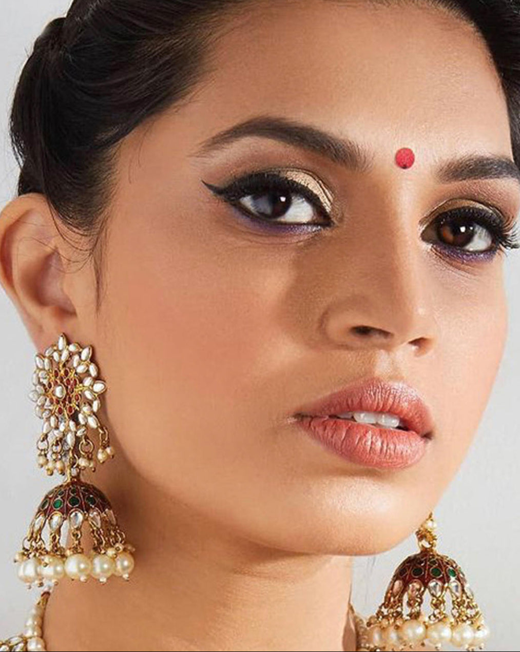 A close-up image of a woman's face, focusing on her wearing gold-plated enameled jhumka earrings embellished with maroon kundan stones and pearls.