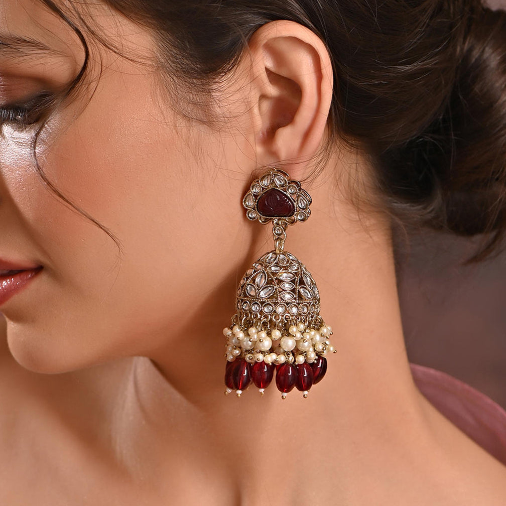 A woman wearing merlot burgundy jhumka earrings with kundan stones, beige pearls, and maroon beads.