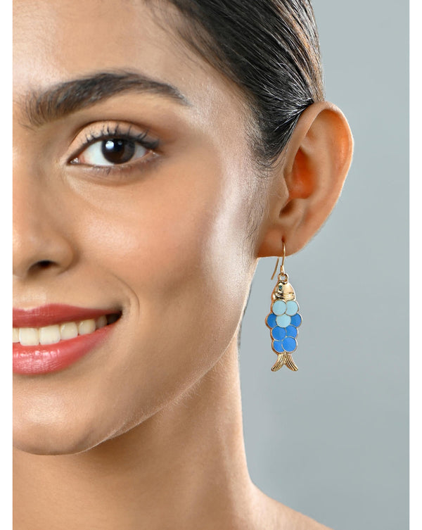 A woman wearing gold-toned drop earrings with fish-shaped charms in navy blue and white.
